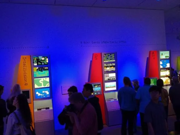 Visitors in front of tall kiosks showing 8-bit game screenshots, with the Intellivision kiosk on the left