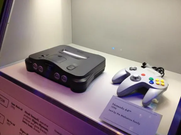 A Nintendo 64 console and controller in a museum case beside its label
