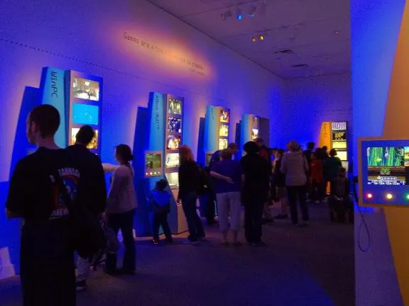 Crowds moving between lit game kiosks in a blue-lit gallery