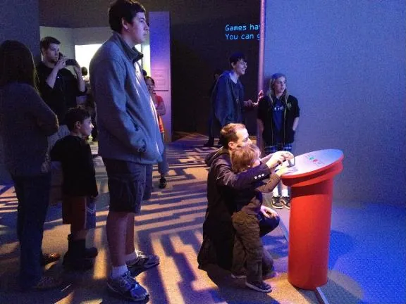 A parent kneels with a small child at a red game pedestal while a line of visitors waits